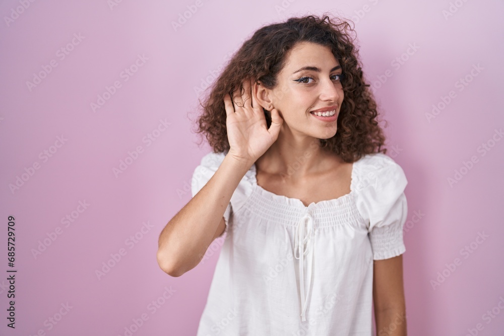 Hispanic woman with curly hair standing over pink background smiling with hand over ear listening an hearing to rumor or gossip. deafness concept.