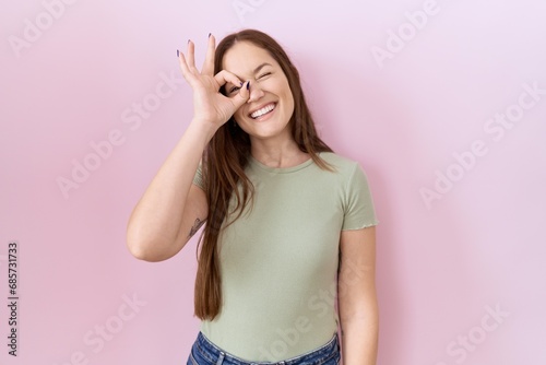 Beautiful brunette woman standing over pink background doing ok gesture with hand smiling  eye looking through fingers with happy face.