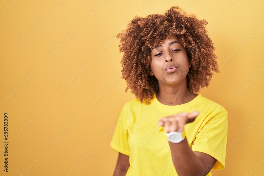 Young hispanic woman with curly hair standing over yellow background looking at the camera blowing a kiss with hand on air being lovely and sexy. love expression.