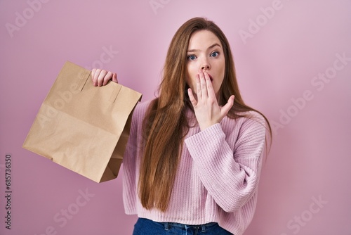 Young caucasian woman holding shopping bag and credit card covering mouth with hand, shocked and afraid for mistake. surprised expression