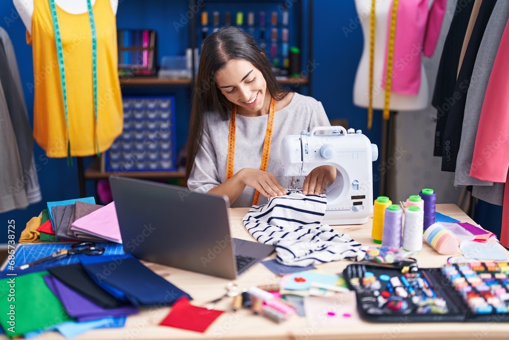 Young beautiful hispanic woman tailor using sewing machine and laptop at clothing factory