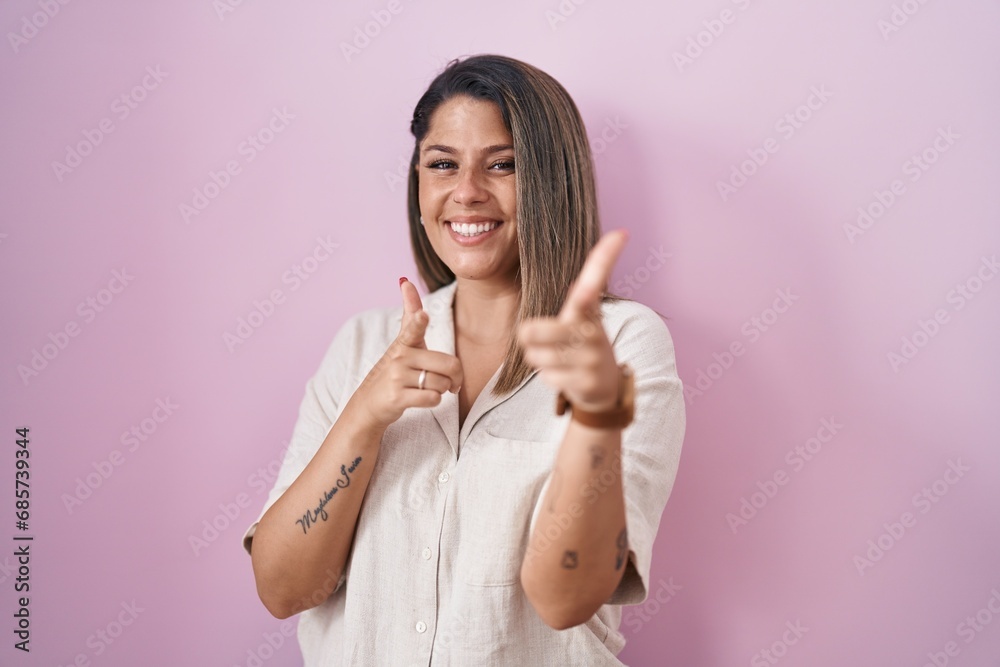 Blonde woman standing over pink background pointing fingers to camera with happy and funny face. good energy and vibes. Blonde woman standing over pink background pointing fingers to camera with happy and funny face. good energy and vibes.