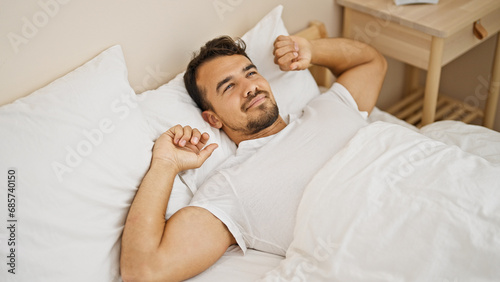 Young hispanic man waking up stretching arms at bedroom