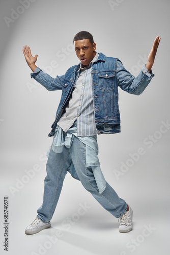 handsome emotional african american man in denim outfit posing on white backdrop, fashion concept