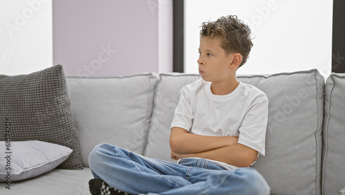 Adorable blond boy sitting in a cozy living room, arms crossed in an upset expression, exuding a serious vibe on the comfortable sofa at home.