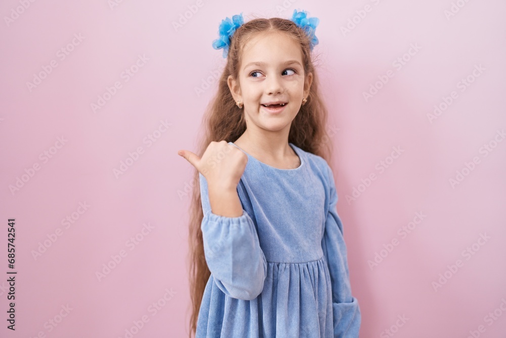 Young little girl standing over pink background smiling with happy face looking and pointing to the side with thumb up. Young little girl standing over pink background smiling with happy face looking and pointing to the side with thumb up.