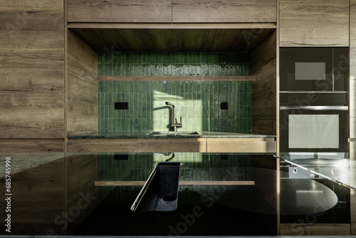 Ceramic hob on an island in front of the sink in a designer kitchen with a green marble countertop, tiles on the backsplash, black taps and wooden furniture