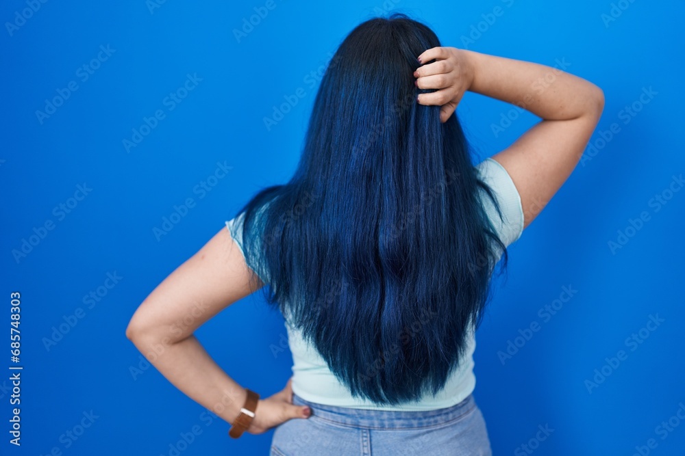 Young modern girl with blue hair standing over blue background backwards thinking about doubt with hand on head Young modern girl with blue hair standing over blue background backwards thinking about doubt with hand on head