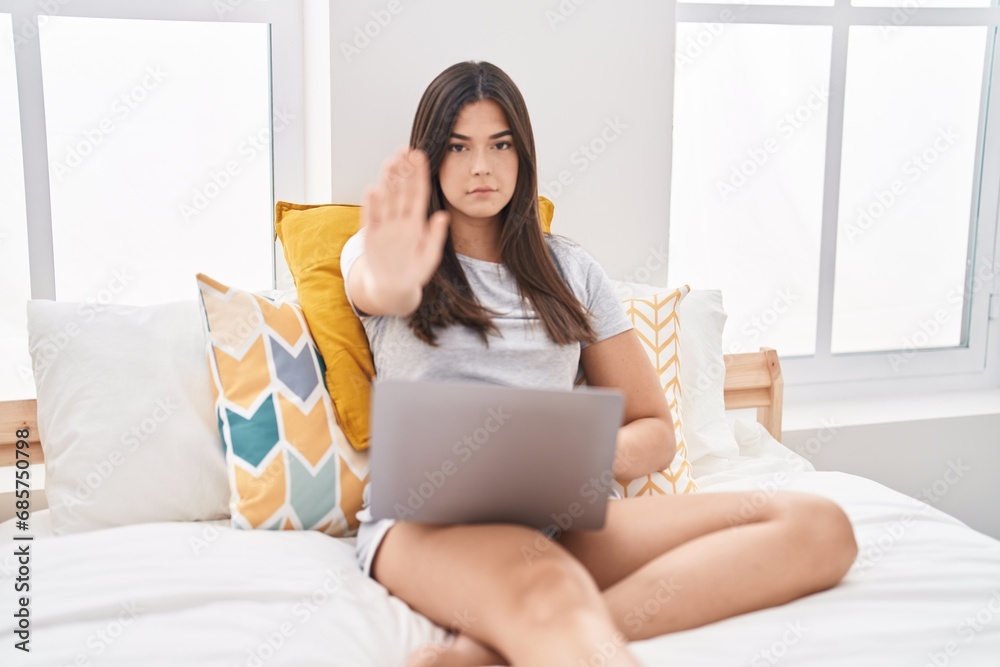 Hispanic woman using computer laptop on the bed with open hand doing stop sign with serious and confident expression, defense gesture Hispanic woman using computer laptop on the bed with open hand doing stop sign with serious and confident expression, defense gesture
