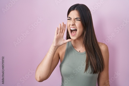 Hispanic woman standing over pink background shouting and screaming loud to side with hand on mouth. communication concept. photo