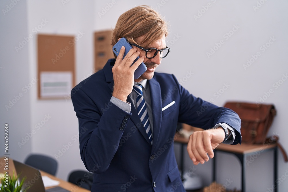 Young blond man business worker talking on smartphone looking watch at office Young blond man business worker talking on smartphone looking watch at office