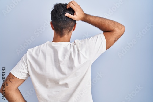 Hispanic man standing over blue background backwards thinking about doubt with hand on head photo