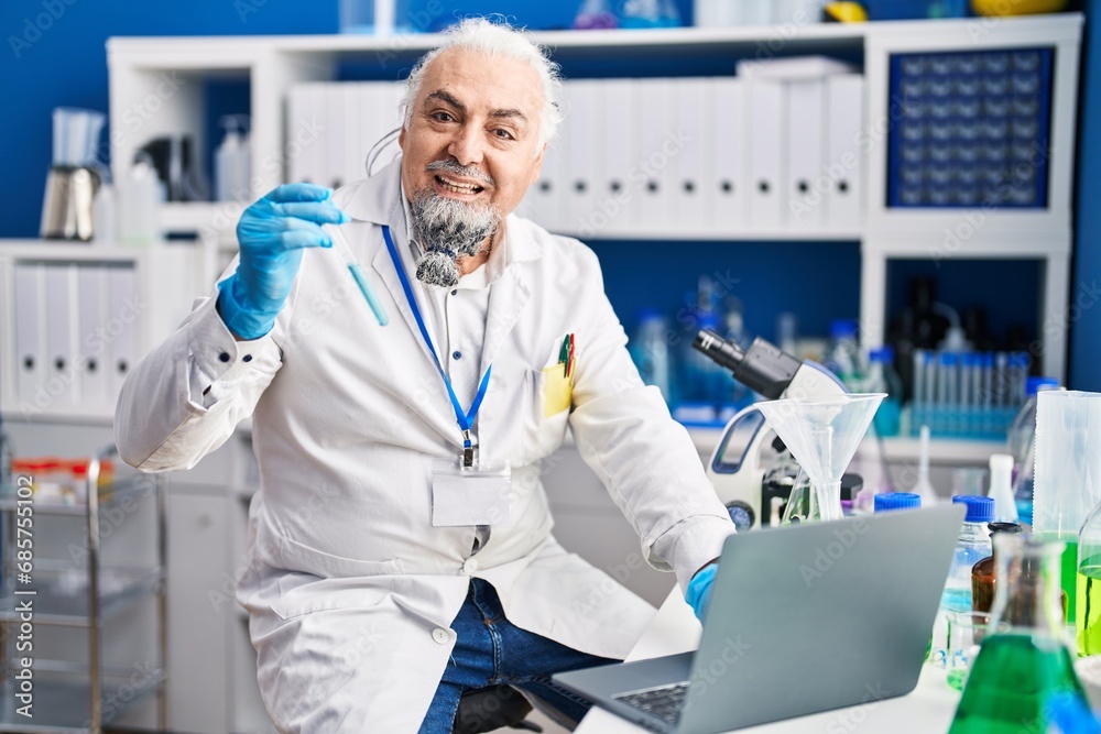 Middle age grey-haired man scientist using laptop holding test tube at laboratory