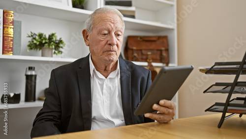 Elegant senior man working relaxed at office, a portrait of success, boss at work using touchpad on laptop at business desk. mature male with grey hair in indoor workplace.