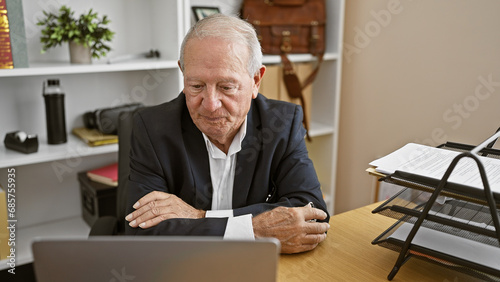 Focused and successful senior man boss sitting at the office desk, arms crossed in confident gesture, seriously looking at laptop screen, immersed in his business work