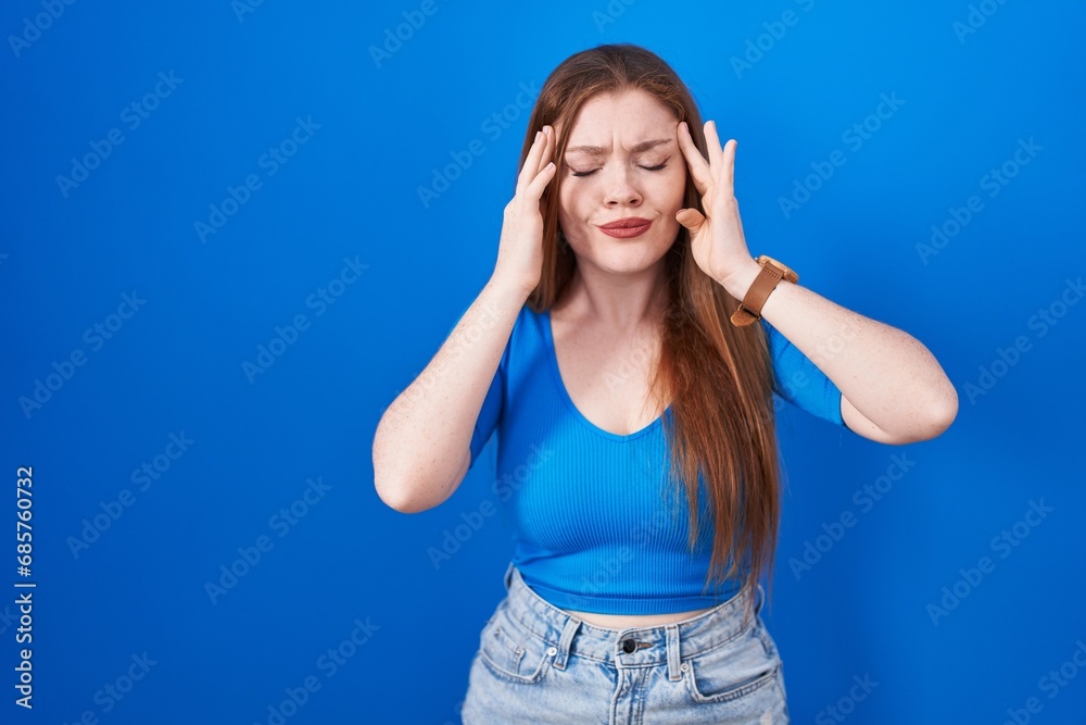 Redhead woman standing over blue background with hand on head, headache because stress. suffering migraine.