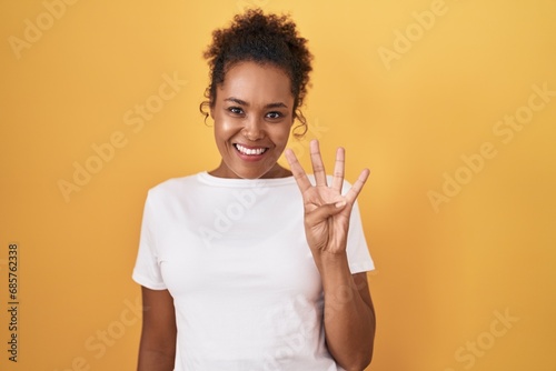 Young hispanic woman with curly hair standing over yellow background showing and pointing up with fingers number four while smiling confident and happy.