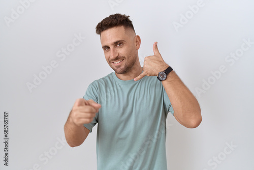 Hispanic man with beard standing over white background smiling doing talking on the telephone gesture and pointing to you. call me.