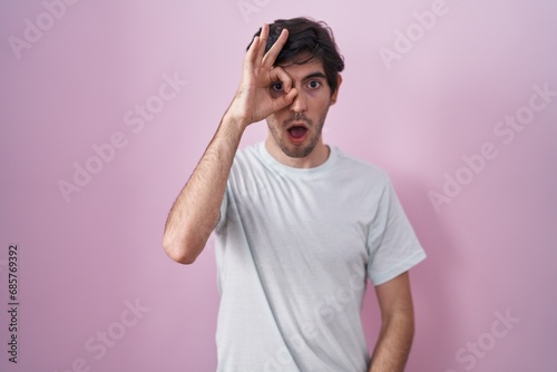 Young hispanic man standing over pink background doing ok gesture shocked with surprised face, eye looking through fingers. unbelieving expression.