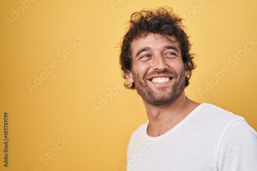 Young hispanic man smiling confident looking to the side over isolated yellow background