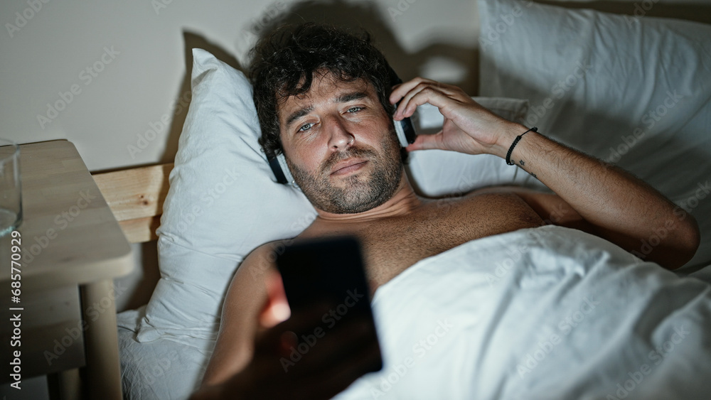 Fototapeta premium Young hispanic man watching video on smartphone lying on bed at bedroom