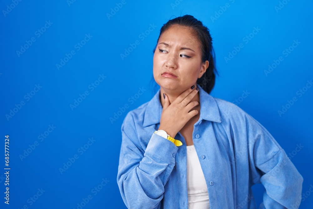Asian young woman standing over blue background touching painful neck, sore throat for flu, clod and infection Asian young woman standing over blue background touching painful neck, sore throat for flu, clod and infection