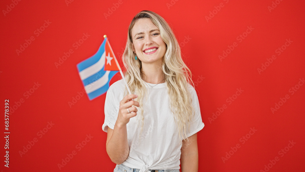 Young blonde woman smiling confident holding cuba flag over isolated red background Young blonde woman smiling confident holding cuba flag over isolated red background