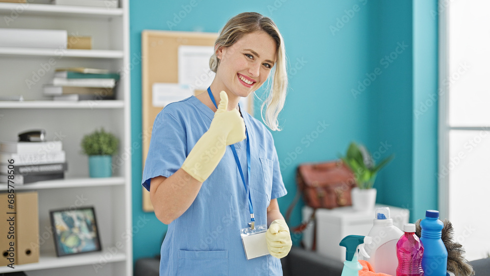 Young blonde woman professional cleaner holding products doing thumb up gesture at the office