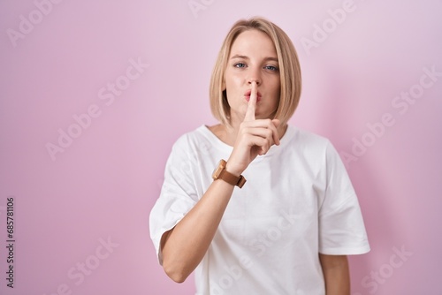 Young caucasian woman standing over pink background asking to be quiet with finger on lips. silence and secret concept.