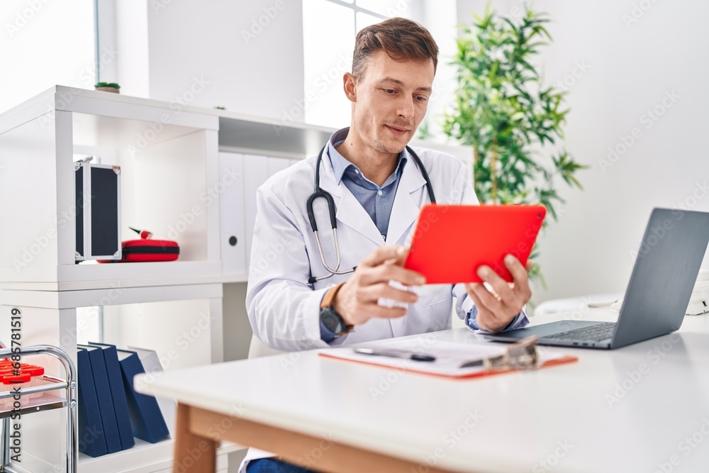 Young man doctor using touchpad sitting on table at clinic Young man doctor using touchpad sitting on table at clinic