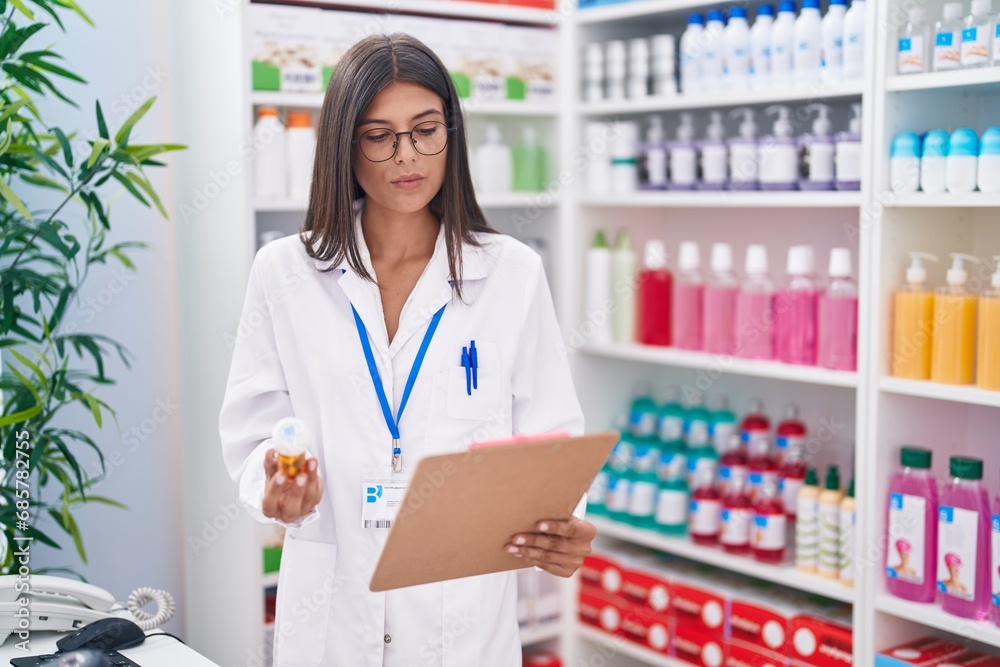 Young beautiful hispanic woman pharmacist holding pills bottle reading document at pharmacy Young beautiful hispanic woman pharmacist holding pills bottle reading document at pharmacy