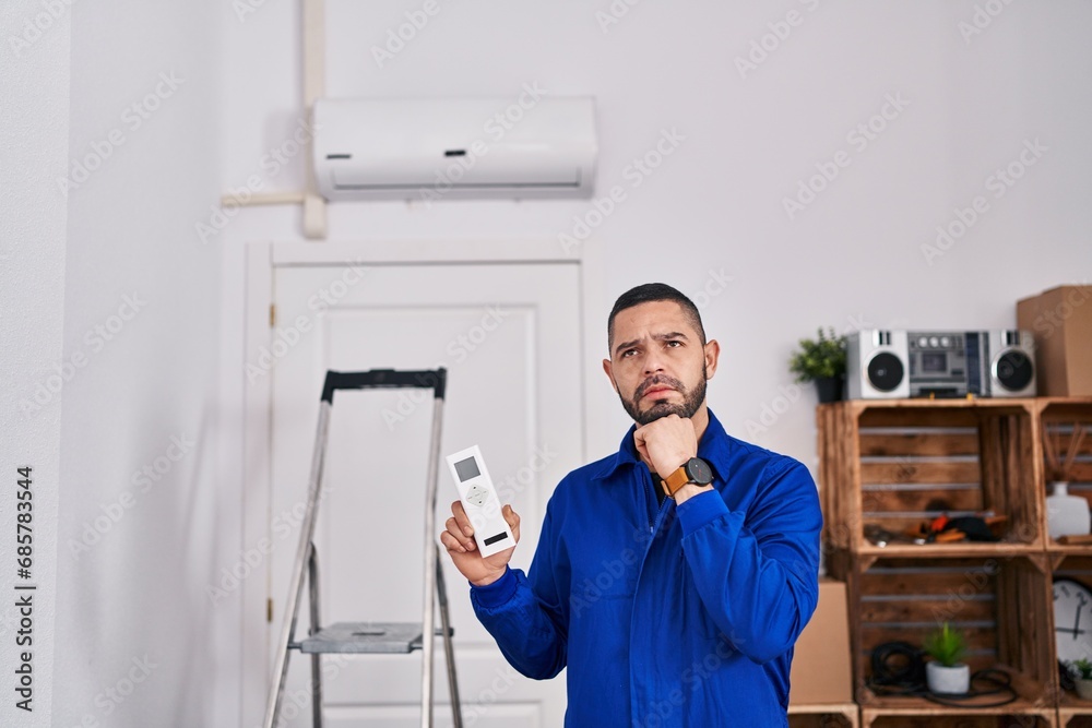 Hispanic repairman working with air conditioner serious face thinking about question with hand on chin, thoughtful about confusing idea