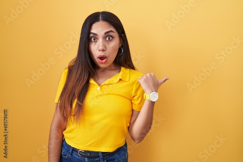 Young arab woman standing over yellow background surprised pointing with hand finger to the side  open mouth amazed expression.