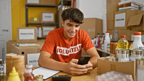 Young hispanic teenager volunteer sitting on table using smartphone smiling at charity center