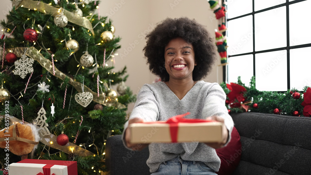 African american woman holding gift sitting on sofa by christmas tree at home