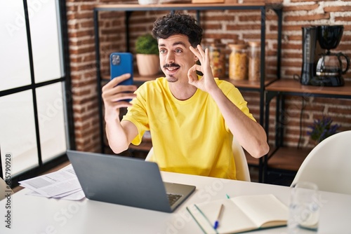 Hispanic man doing video call with smartphone doing ok sign with fingers, smiling friendly gesturing excellent symbol