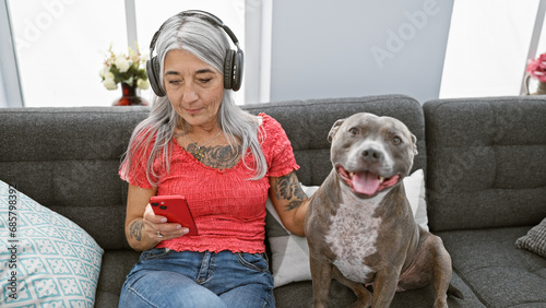 Grey-haired middle age woman sitting on living room sofa, absorbing her song's melody via gadget, serene dog by her side, sharing the audio magic indoors!