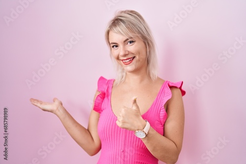 Young caucasian woman standing over pink background showing palm hand and doing ok gesture with thumbs up, smiling happy and cheerful