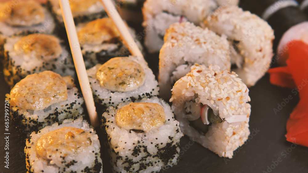 Chopsticks take sushi from a set of sushi rolls. Fresh rolls in a Japanese restaurant. Oriental cuisine. Close-up. A set of delicious Japanese sushi rolls on a stone blackboard.