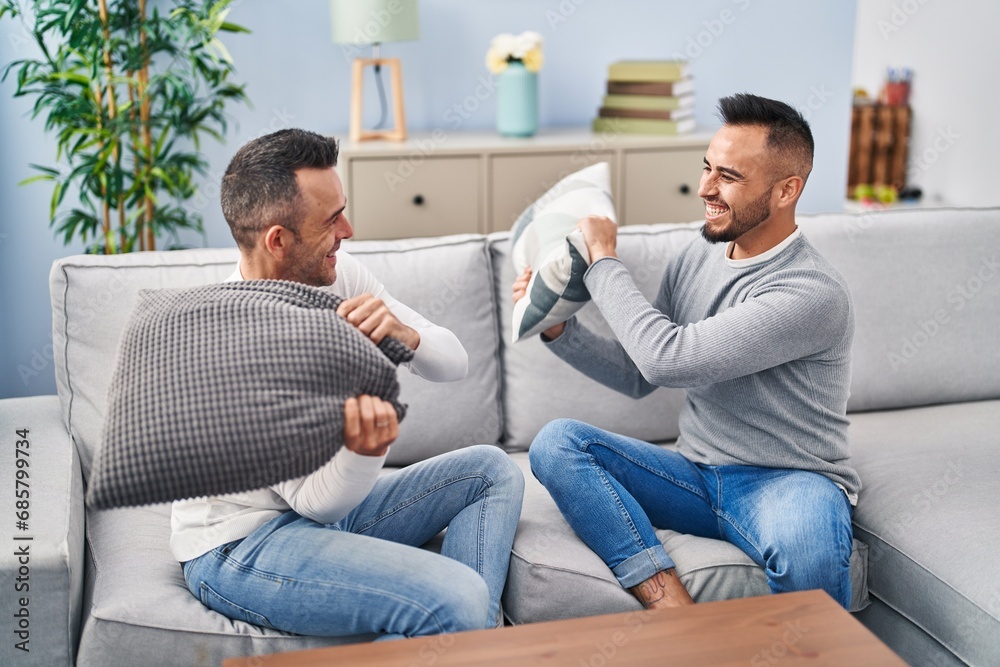 Two men couple fighting with cushion sitting on sofa at home