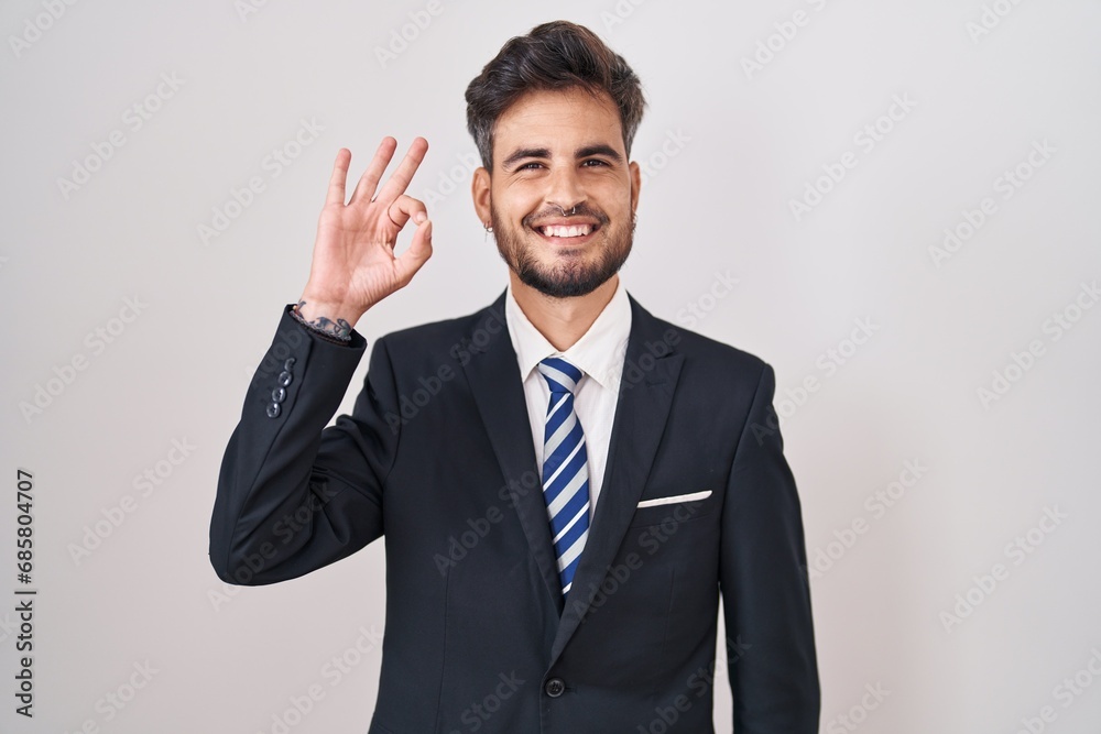 Young hispanic man with tattoos wearing business suit and tie smiling positive doing ok sign with hand and fingers. successful expression. Young hispanic man with tattoos wearing business suit and tie smiling positive doing ok sign with hand and fingers. successful expression.