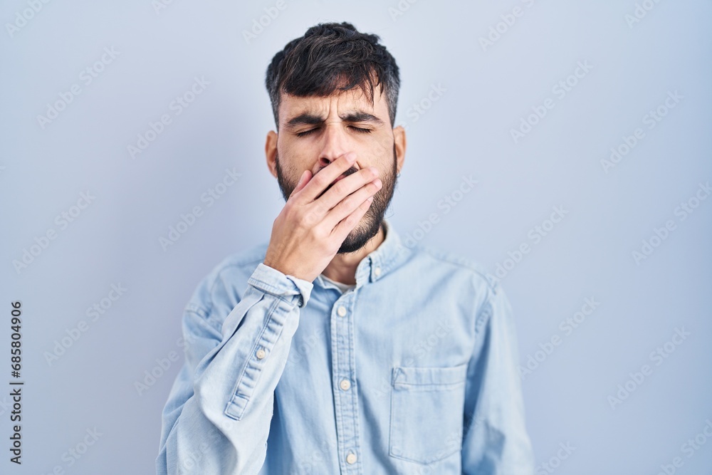 Young hispanic man with beard standing over blue background bored yawning tired covering mouth with hand. restless and sleepiness. Young hispanic man with beard standing over blue background bored yawning tired covering mouth with hand. restless and sleepiness.