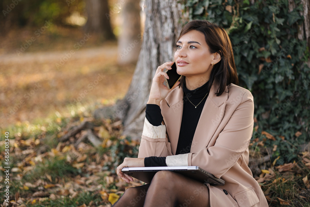 Portrait of attractive female sitting in the park and speak by the phone. Happy woman spent time on open air Portrait of attractive female sitting in the park and speak by the phone. Happy woman spent time on open air