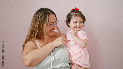 Mother and daughter, happily pointing and smiling together, over a lovely pink isolated background in a casual, confident display of maternal love and joyful family lifestyle.