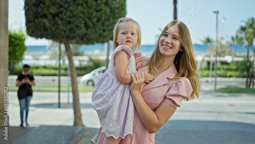 Joyful, confident mother smiling, holding her cheerful daughter in her arms, standing together on a sunny, urban street