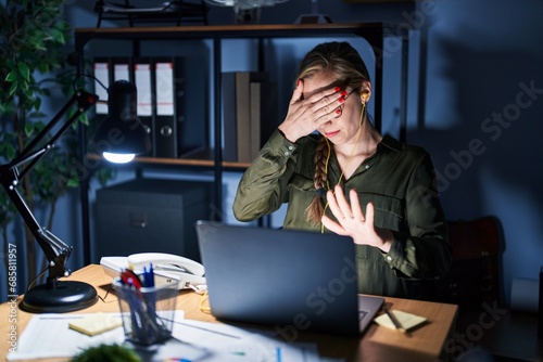 Young blonde woman working at the office at night covering eyes with hands and doing stop gesture with sad and fear expression. embarrassed and negative concept.