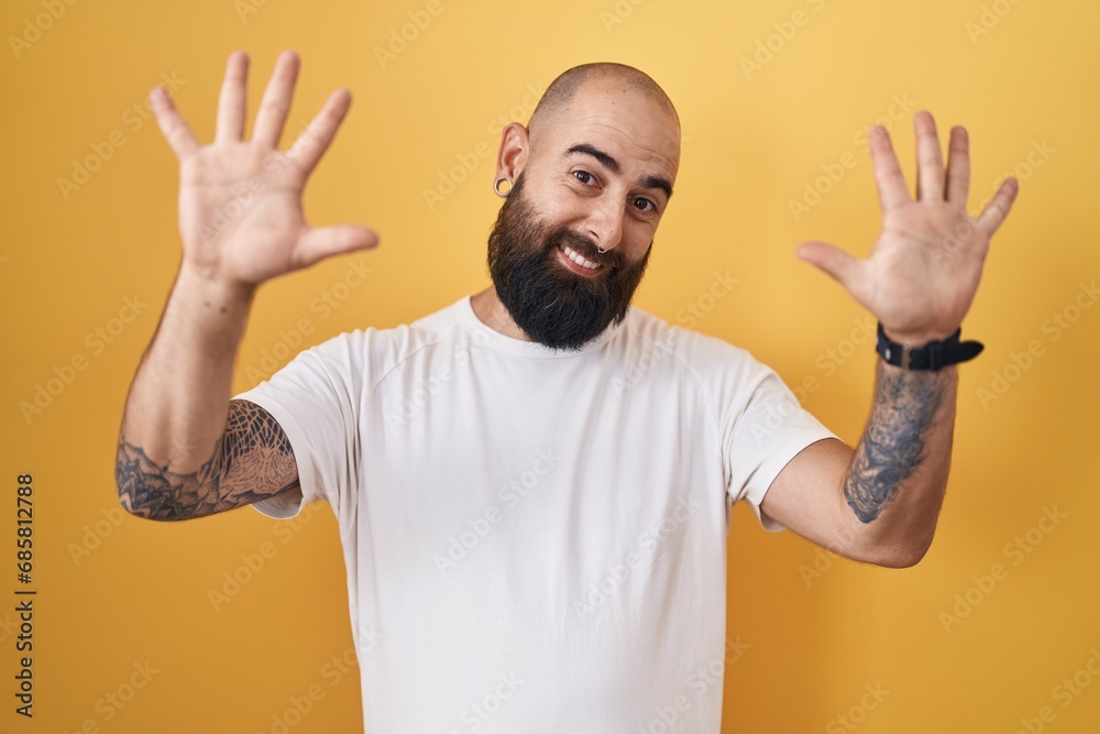 Fototapeta premium Young hispanic man with beard and tattoos standing over yellow background showing and pointing up with fingers number ten while smiling confident and happy.