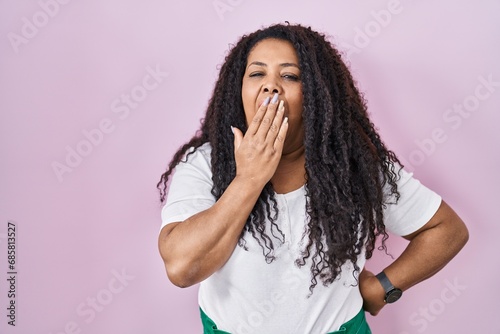 Plus size hispanic woman standing over pink background bored yawning tired covering mouth with hand. restless and sleepiness.