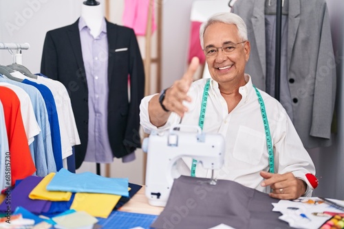 Middle age man with grey hair dressmaker using sewing machine smiling friendly offering handshake as greeting and welcoming. successful business.
