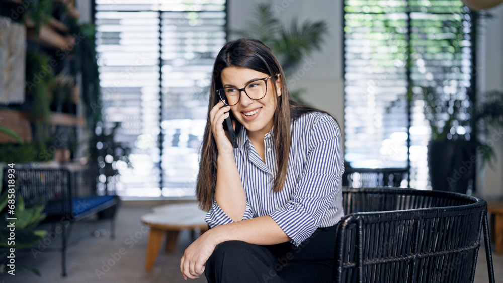Fototapeta premium Young beautiful hispanic woman speaking on the phone sitting on a chair at the office